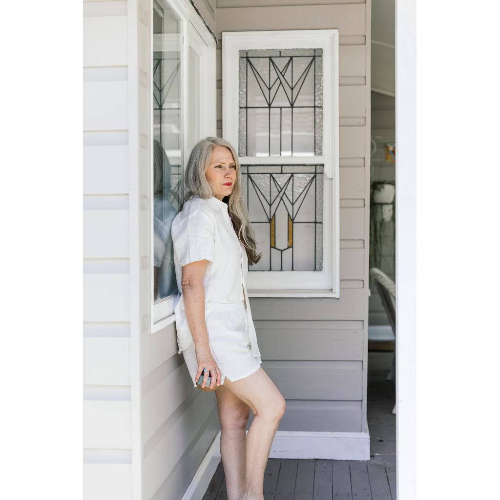 French flax linen unisex boxer shorts, loose, comfy and playful for men and women of all shapes and sizes - Available in 5 colours: White, Pinstripe, Natural, Navy & Charcoal – White boxer shorts pictured on a woman leaning on the side of a weatherboard house - available at LOVINLIFE Co Byron Bay for all your gifts, candles and interior decorating needs