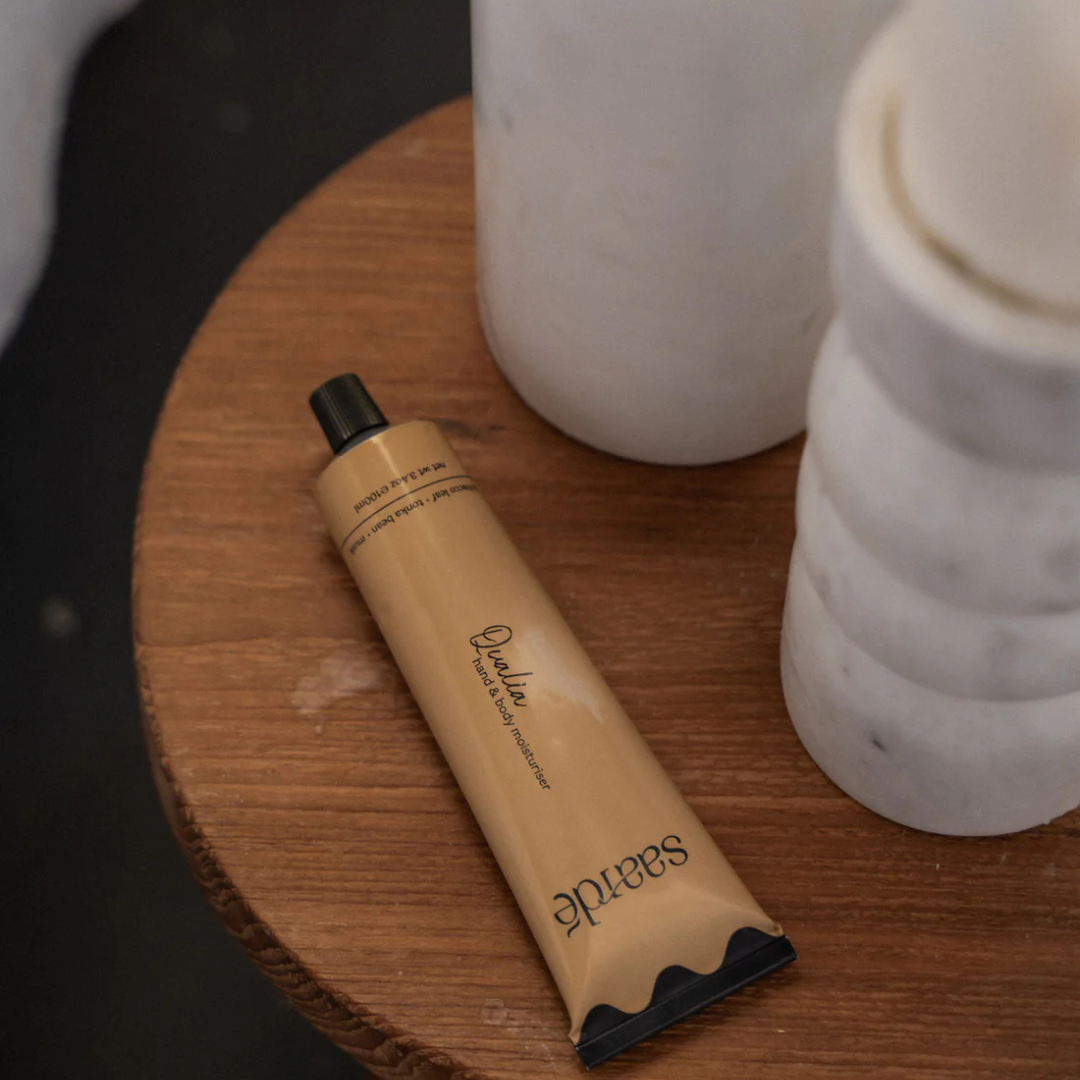 Hand & Body Moisturiser by Saarde - Qualia pictured from above lying on a coffee table next to a candle holder - Stocked at LOVINLIFE Co Byron Bay for all your gifts, candles and interior decorating needs