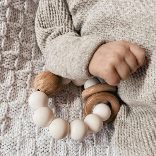 Child's hand holding a wooden teething ring on a textured surface. White Silicone Ring teething toy with natural wood bead and rings - Available at LOVINLIFE Co Byron Bay