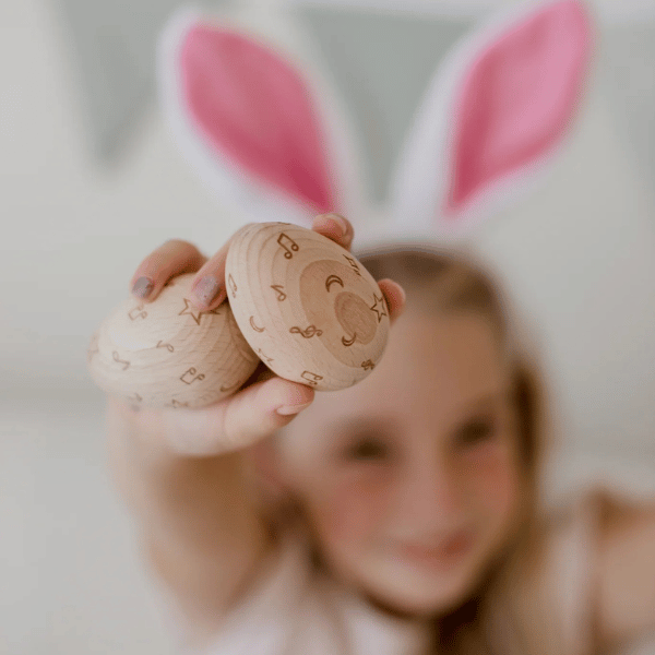 Child with bunny ears blurred in background holding 2 x wooden egg shakers by Love Note Co made with FSC certified Beechwood. All  instruments by Love Note Co are handmade items, non-toxic, eco-friendly and sustainably sourced. Stocked at LOVINLIFE Co Byron Bay for all your gifts, candles and interior decorating needs