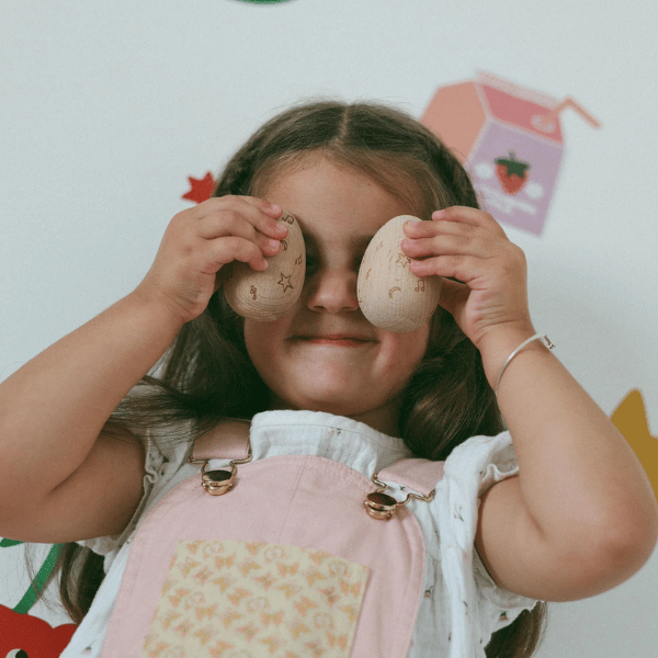 Child holding 2 x wooden egg shakers up in front of her eyes. Egg shakers are by Love Note Co made with FSC certified Beechwood. All  instruments by Love Note Co are handmade items, non-toxic, eco-friendly and sustainably sourced. Stocked at LOVINLIFE Co Byron Bay for all your gifts, candles and interior decorating needs