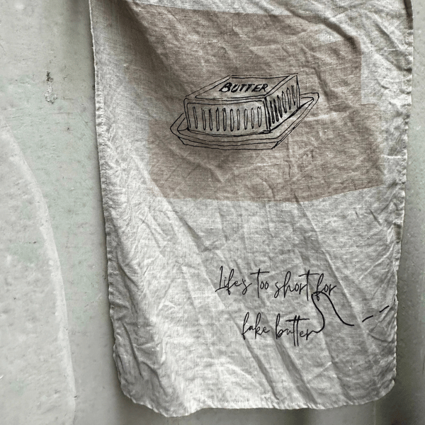 French Linen Tea Towel pictured hanging to show a sketch of a butter dish and the words "Life's too short for fake butter..." printed underneath. Designed in Australia, Heaven In Earth, available at LOVINLIFE Co Byron Bay