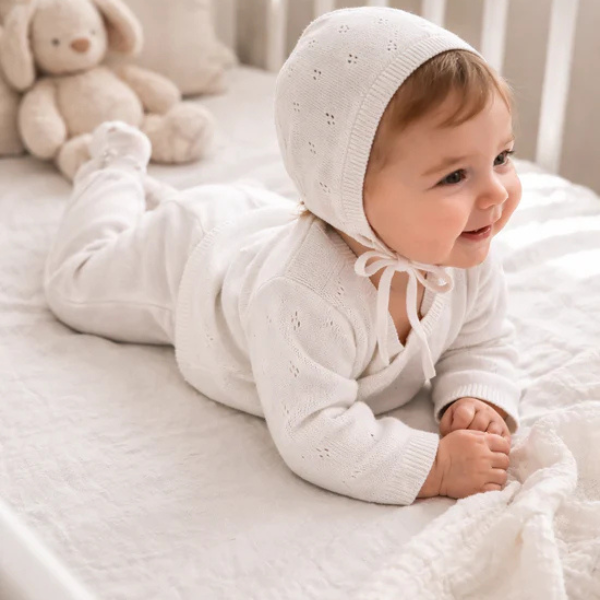 Baby showcasing an 100% organic cotton 4-piece Heirloom Knit Baby Gift Set in white with a plush toy in the background. Set by Aster & Oak and Stocked at LOVINLIFE Co Byron Bay for all your gifts, candles and interior decorating needs