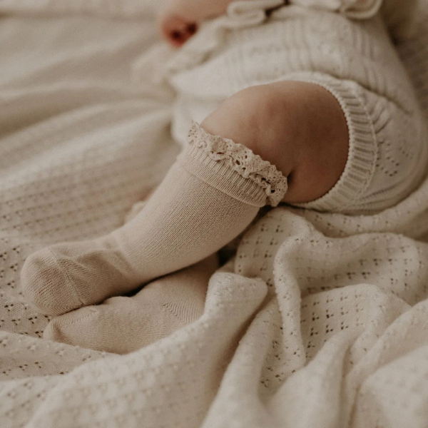 Close-up of a newborn baby's legs wearing beige socks with lace trim on a soft, organic cotton textured Knit Heirloom Baby Blanket in Cream. Stocked at LOVINLIFE Co Byron Bay for all your gifts, candles and interior decorating needs