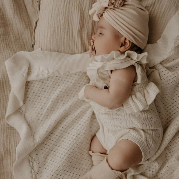 Baby in a beige outfit and turban lying on an organic cotton textured Knit Heirloom Baby Blanket in Cream. Stocked at LOVINLIFE Co Byron Bay for all your gifts, candles and interior decorating needs