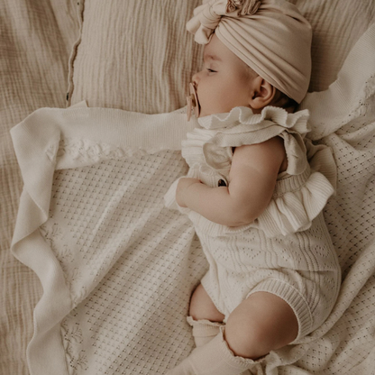 Baby in a beige outfit and turban lying on an organic cotton textured Knit Heirloom Baby Blanket in Cream. Stocked at LOVINLIFE Co Byron Bay for all your gifts, candles and interior decorating needs