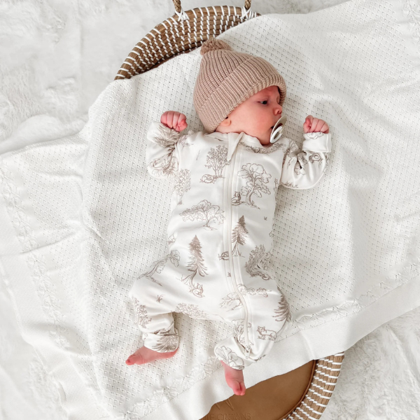 Newborn baby in a white patterned outfit and sand coloured beanie lying on an organic cotton textured Knit Heirloom Baby Blanket in Cream. Stocked at LOVINLIFE Co Byron Bay for all your gifts, candles and interior decorating needs