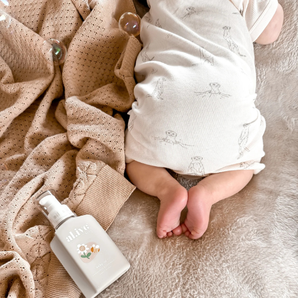 Baby's legs and feet with a bottle of baby lotion crawling on an organic cotton textured Knit Heirloom Baby Blanket in Sand, wearing a brown outfit. Stocked at LOVINLIFE Co Byron Bay for all your gifts, candles and interior decorating needs