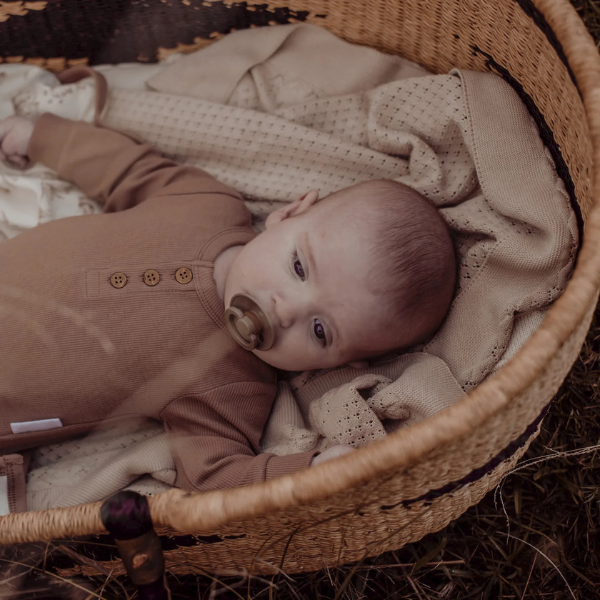 Baby lying in a wicker basket with an organic cotton textured Knit Heirloom Baby Blanket in Sand, wearing a brown outfit. Stocked at LOVINLIFE Co Byron Bay for all your gifts, candles and interior decorating needs