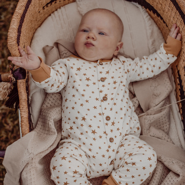 Baby in a star-patterned onesie lying in a wicker crib on top of an organic cotton textured Knit Heirloom Baby Blanket in Sand, wearing a brown outfit. Stocked at LOVINLIFE Co Byron Bay for all your gifts, candles and interior decorating needs