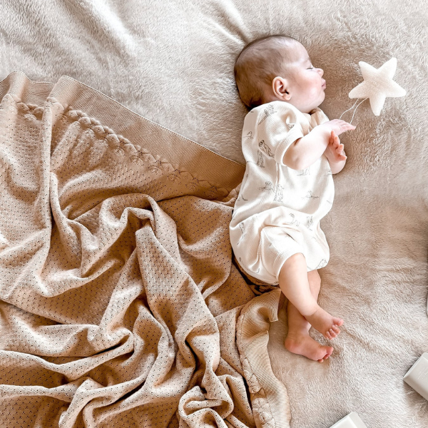 Baby lying on a soft surface with a star-shaped toy, surrounded by an organic cotton textured Knit Heirloom Baby Blanket in Sand. Stocked at LOVINLIFE Co Byron Bay for all your gifts, candles and interior decorating needs