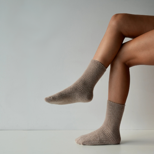 Taupe coloured socks on a person’s feet, side view of them sitting with their legs crossed and one foot in the air, on a plain background. Luxe Cashmere Blend Socks by Number Thirty One made with a premium blend of cashmere and wool. Unisex & One size. Stocked at LOVINLIFE Co Byron Bay for all your gifts, candles and interior decorating needs