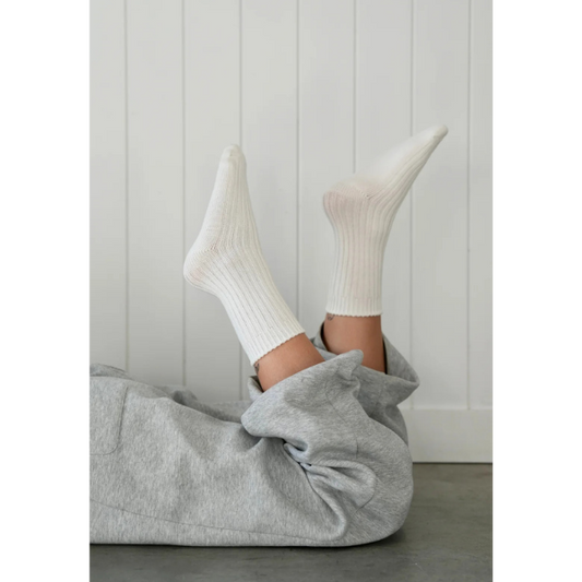 Person wearing white socks and grey trackpants lying on their stomach on the floor with feet in the air in front of a white panelled wall. Luxe Cotton Socks by Number Thirty One made with a soft cotton, wool, and spandex blend. Unisex & One size. Stocked at LOVINLIFE Co Byron Bay for all your gifts, candles and interior decorating needs