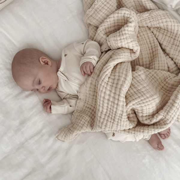 Baby wrapped in an organic muslin cotton baby blanket in a sand colour on a white surface. Stocked at LOVINLIFE Co Byron Bay for all your gifts, candles and interior decorating needs
