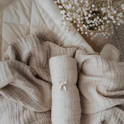 Flat lay of an organic muslin cotton baby blanket in sand rolled up on top of another lighter coloured baby blanket on a bed. Stocked at LOVINLIFE Co Byron Bay for all your gifts, candles and interior decorating needs