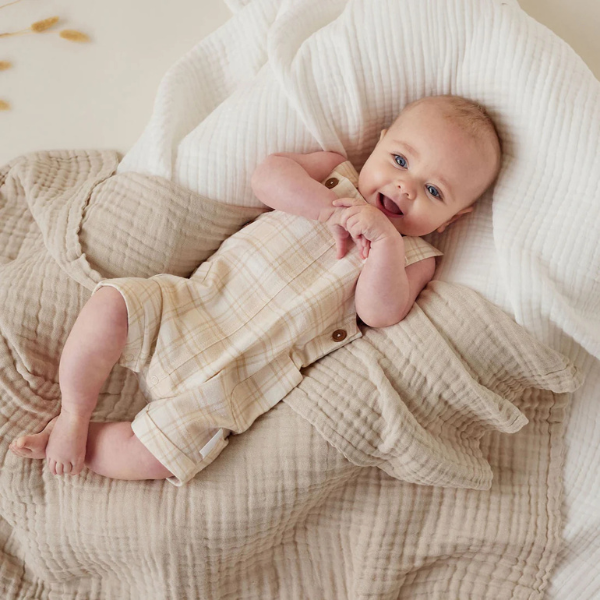 Baby in a beige outfit lying on an organic muslin cotton baby blanket in sand. Stocked at LOVINLIFE Co Byron Bay for all your gifts, candles and interior decorating needs