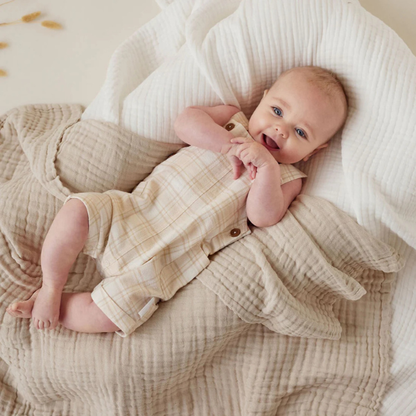 Baby in a beige outfit lying on an organic muslin cotton baby blanket in sand. Stocked at LOVINLIFE Co Byron Bay for all your gifts, candles and interior decorating needs