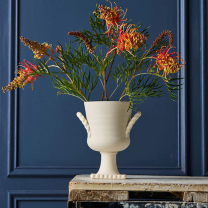 White vase with greenery and Australian Native Flowers against a blue wall. Vase Vintage Trophy Life - Florence. Part of the Robert Gordon Collection stocked at LOVINLIFE Co Byron Bay for all your gifts, candles, homewares and interior decorating needs