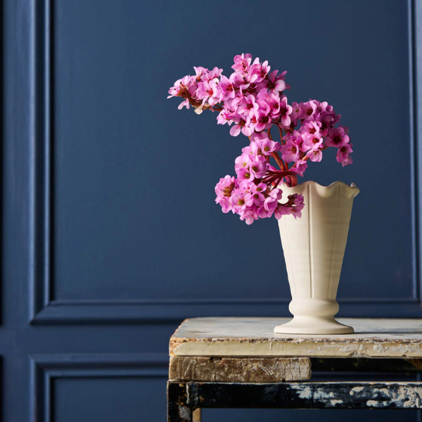 White vase with pink flowers on a wooden table against a blue wall. Vase Vintage Trophy Life - Violet. Part of the Robert Gordon Collection stocked at LOVINLIFE Co Byron Bay for all your gifts, candles, homewares and interior decorating needs