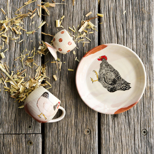 Ceramic cup and saucer with chicken design on a wooden surface with hay. A Henny Penny Chickens design, part of the Robert Gordon Collection stocked at LOVINLIFE Co Byron Bay for all your gifts, candles, homewares and interior decorating needs