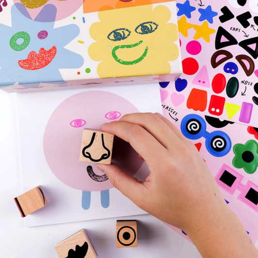 Child's hand using a stamp with a nose on a piece of paper surrounded by colourful stamps and stickers. From Wooden Stamp Kit - Cool Faces - Stocked at LOVINLIFE Co Byron Bay for all your gifts, candles, homewares and interior decorating needs