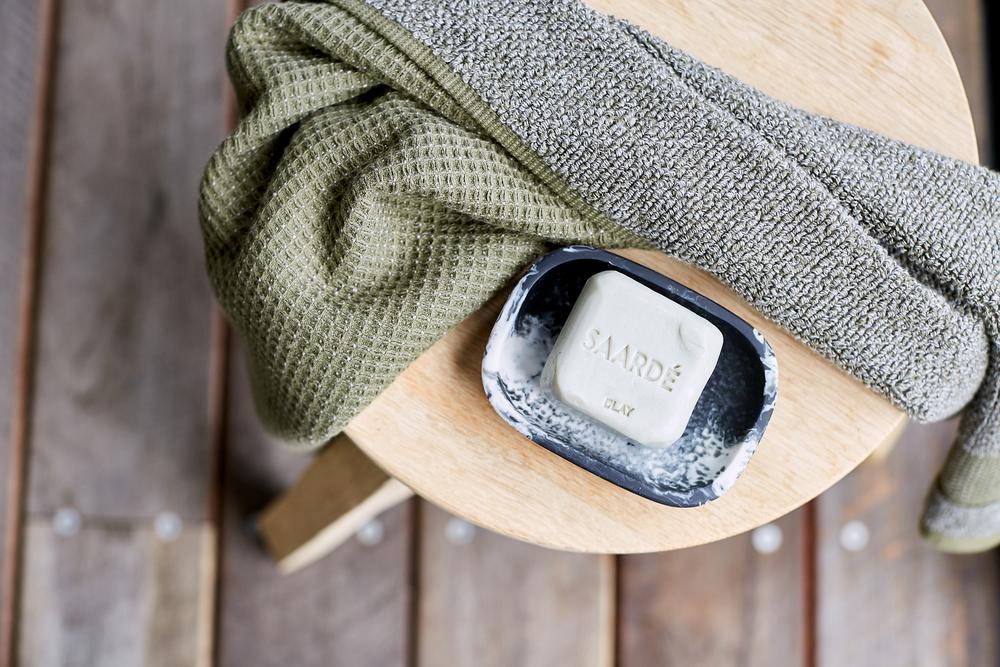 Lovinlife Co ByronBay – Saarde Clay Olive Oil Bar Soap – draws out impurities from blocked pores, beneficial for acne prone or oily skin - unscented – soap bar pictured as flay lay in black resin soap dish on a wooden stool with an olive towel - available at LOVINLIFE Co Byron Bay for all your gifts, candles and interior decorating needs